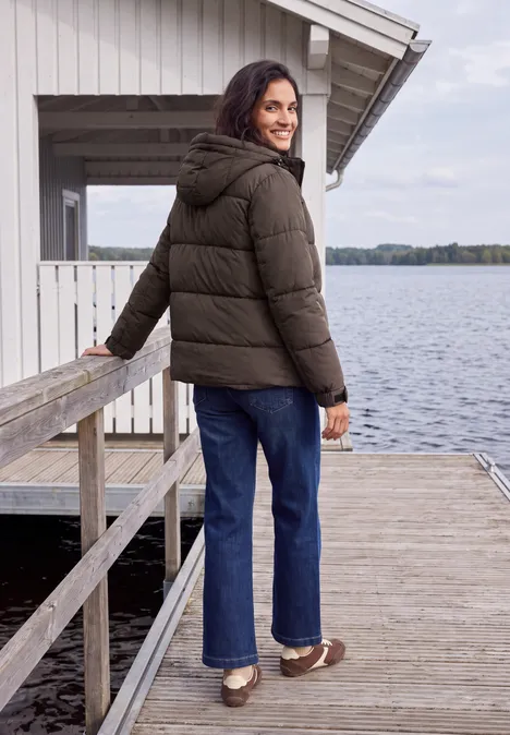 Frau in grüner Daunenjacke steht auf Steg am See und blickt lächelnd zurück.