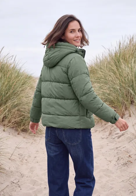 Frau in grüner Steppjacke läuft am Strand zwischen Dünen und blickt lächelnd zurück.
