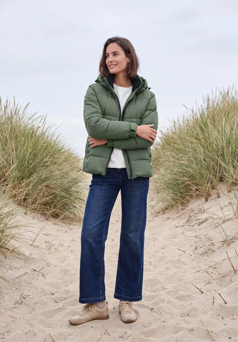 Eine lächelnde Frau in grüner Steppjacke, Jeans und hellen Schuhen steht auf Sanddünen.