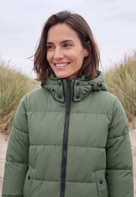 Eine lächelnde Frau in einer grünen Steppjacke mit Kapuze, steht am Strand.