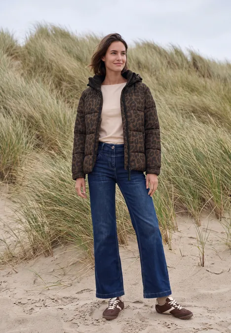 Frau in Steppjacke, Jeans und Sneakern steht auf Sand vor Dünen.