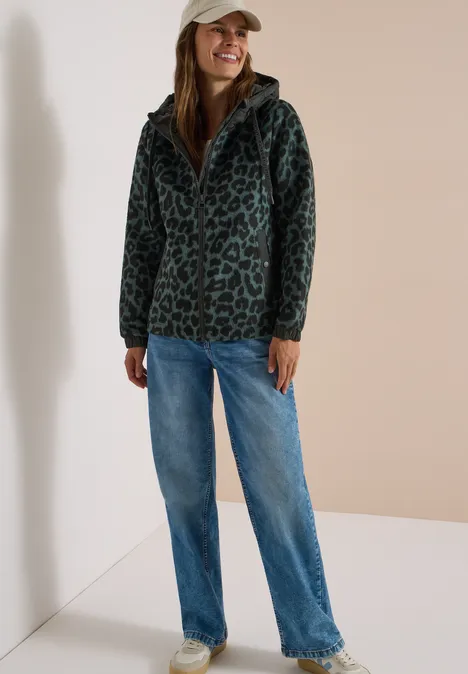 Frau in leo-gepunkteter Fleecejacke, blauen Jeans, weißen Sneakern und Baseballcap.