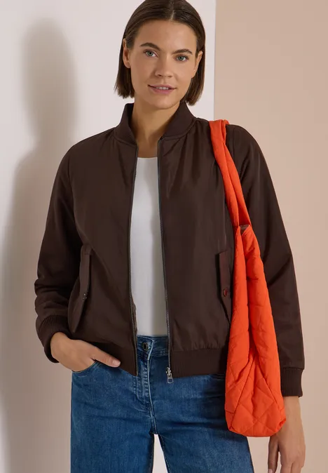 Frau mit weißem T-Shirt, brauner Bomberjacke, blauer Jeans und orangefarbener Stepp-Tasche über der Schulter.