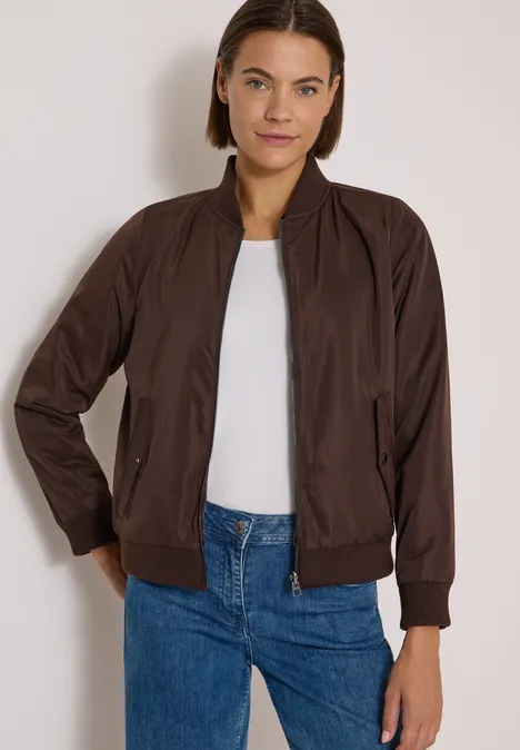 Frau in brauner Bomberjacke, weißem Shirt und blauen Jeans vor hellem Hintergrund.