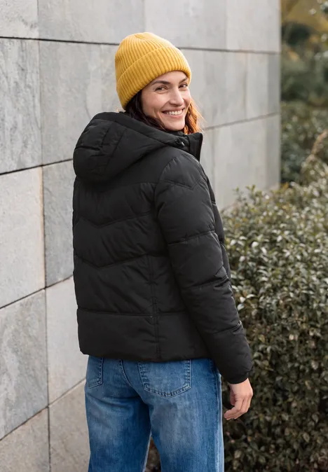 Veste matelassée à capuche avec des détails en sergé Black Eine Person mit gelber Strickmütze und schwarzer Steppjacke steht an einer Wand und blickt lächelnd zurück.