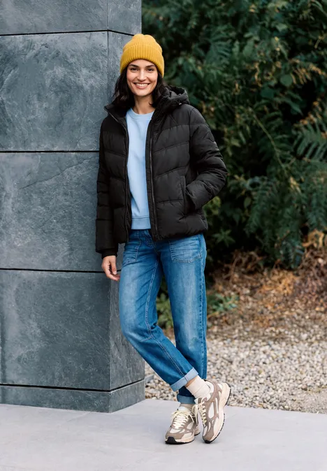 Veste matelassée à capuche avec des détails en sergé Black Eine junge Frau lehnt lächelnd an einer Wand, trägt schwarze Steppjacke, blaue Jeans, gelbe Mütze.