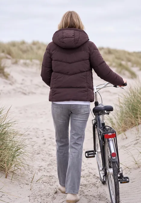 Veste matelassée à capuche avec des détails en sergé berry red Une femme en doudoune pousse un vélo le long d’un sentier de dunes à la plage.