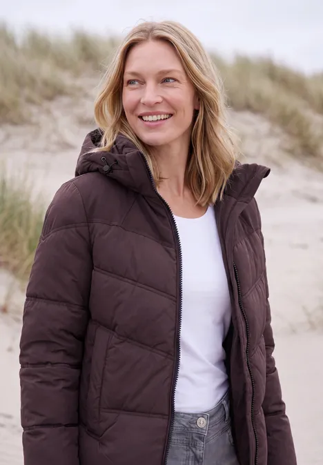 Veste matelassée à capuche avec des détails en sergé berry red Une femme souriante en doudoune marron et t-shirt blanc sur une plage devant des dunes de sable.
