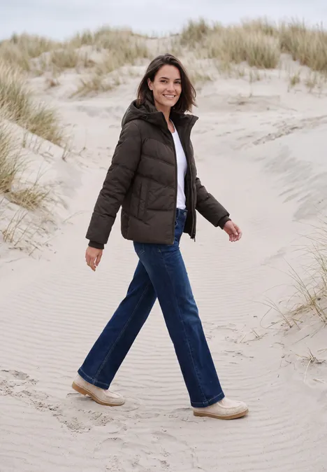 Veste matelassée à capuche avec des détails en sergé chocolate sauce brown Une femme souriante en doudoune et jean marche paisiblement dans les dunes au bord de la mer.