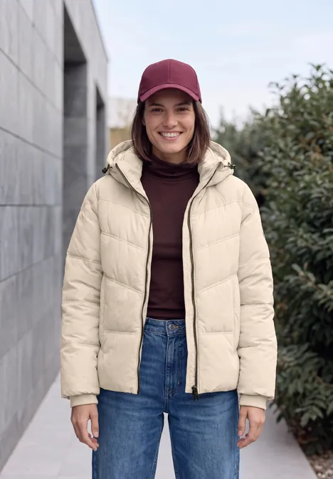 Veste matelassée à capuche avec détails en sergé powder beige Jeune femme souriante en doudoune beige et bonnet bordeaux devant un mur et des arbustes.