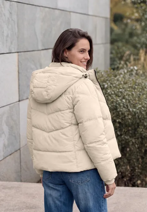 Veste matelassée à capuche avec détails en sergé powder beige Femme en doudoune crème adossée à un mur de pierre, regard souriant par-dessus l'épaule.