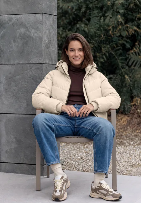Veste matelassée à capuche avec détails en sergé powder beige Jeune femme assise dehors sur une chaise, veste matelassée beige, jean bleu et baskets, sourire détendu.