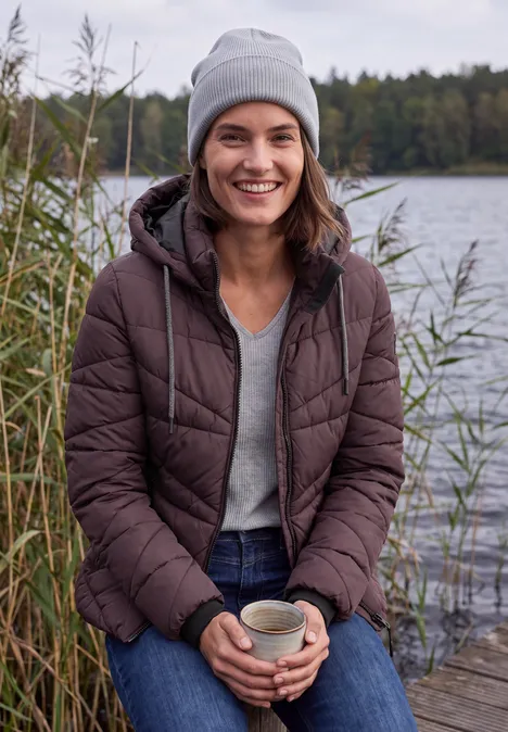 Vrouw in een donzen jas en muts zit lachend op een steiger aan het meer met een mok in haar handen.
