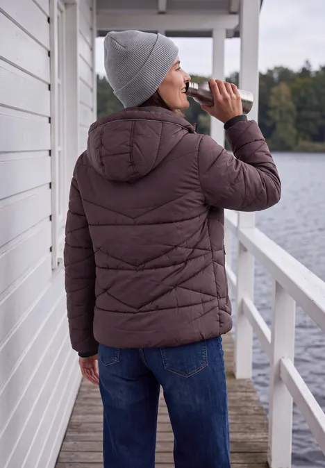 Vrouw in winterjas en muts drinkt uit een fles op een steiger aan het water.