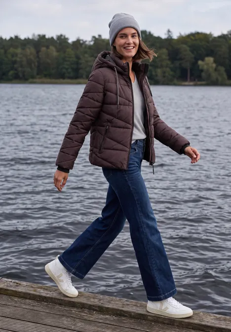 Een lachende vrouw in jeans, winterjas en muts wandelt over een steiger langs het meer.