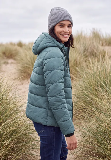 Steppjacke mit Kapuze und Zipper mountain air blue Frau in grüner Steppjacke und grauer Mütze steht in Dünenlandschaft und lächelt über die Schulter.