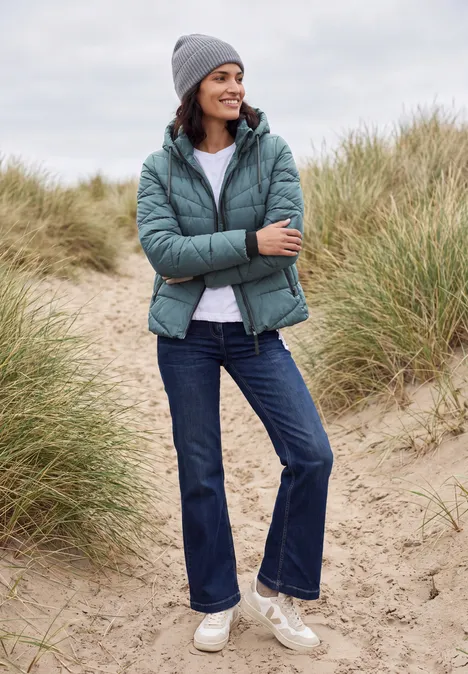Steppjacke mit Kapuze und Zipper mountain air blue Frau in Winterjacke, Jeans, Mütze und Sneakers steht lächelnd zwischen Dünen am Strand