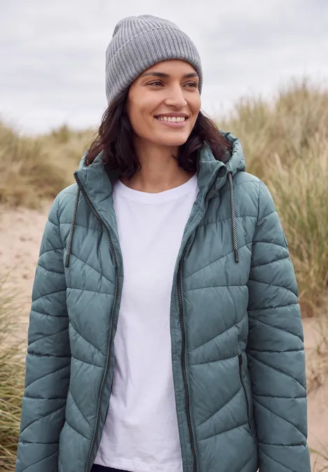 Steppjacke mit Kapuze und Zipper mountain air blue Frau mit grauer Mütze und grüner Steppjacke steht lächelnd in Dünen.