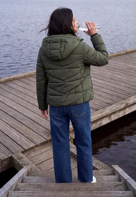 Steppjacke mit Kapuze und Zipper olive night Person mit grüner Steppjacke und blauer Jeans trinkt am Holzsteg, Rückenansicht.
