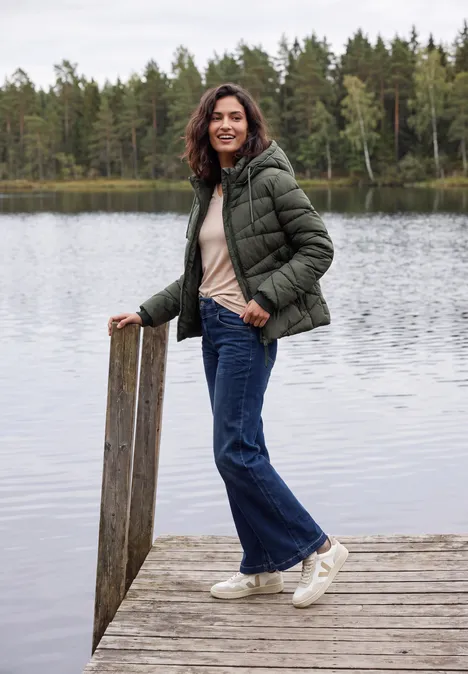 Steppjacke mit Kapuze und Zipper olive night Frau mit grüner Steppjacke, Jeans und weißen Sneakern steht lächelnd auf einem Steg am ruhigen See.