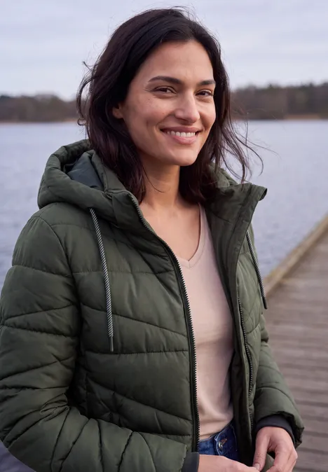 Steppjacke mit Kapuze und Zipper olive night Lächelnde Frau in grüner Steppjacke steht auf einem Steg vor einem See.