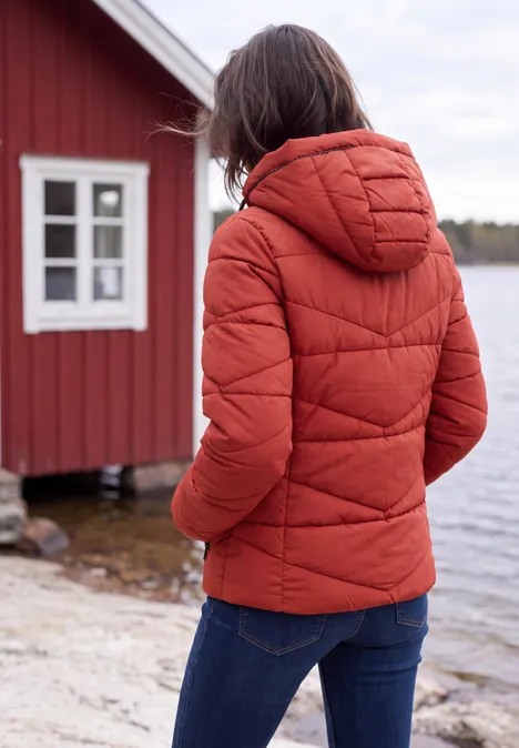 Frau in roter Steppjacke mit Kapuze steht am Ufer vor einem roten Haus.