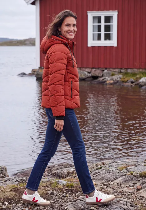 Eine Frau in oranger Steppjacke und Jeans läuft am Ufer vor einer roten Hütte am See.