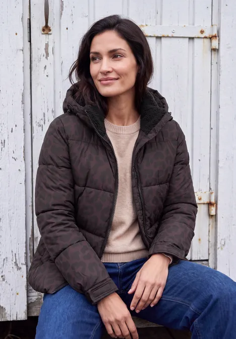 Een vrouw in een donkere, doorgestikte jas zit ontspannen voor een witte houten muur.