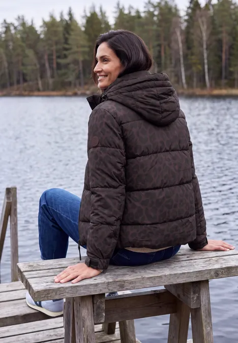 Vrouw zit glimlachend in donsjas op een houten bank bij een meer, omringd door bos en rustig water.