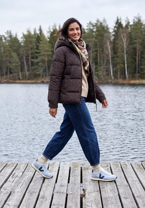 Vrouw in winterjas en sneakers loopt lachend over een steiger bij een meer.