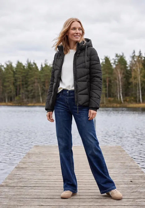 Frau in schwarzer Daunenjacke, weißem Pullover, blauen Jeans und Hausschuhen steht lächelnd auf einem Steg am See.