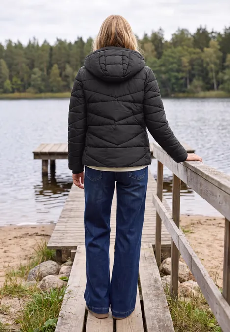 Eine Person mit schwarzer Steppjacke und Jeans steht auf einem Steg am ruhigen Waldsee.