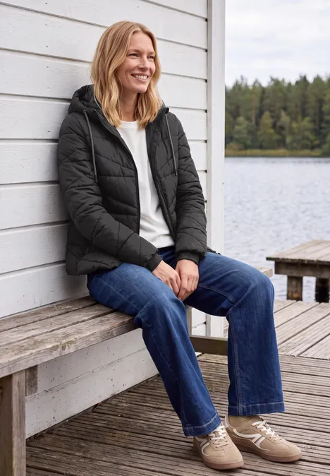 Frau in Jeans und Steppjacke sitzt lächelnd am hölzernen Steg eines Sees.