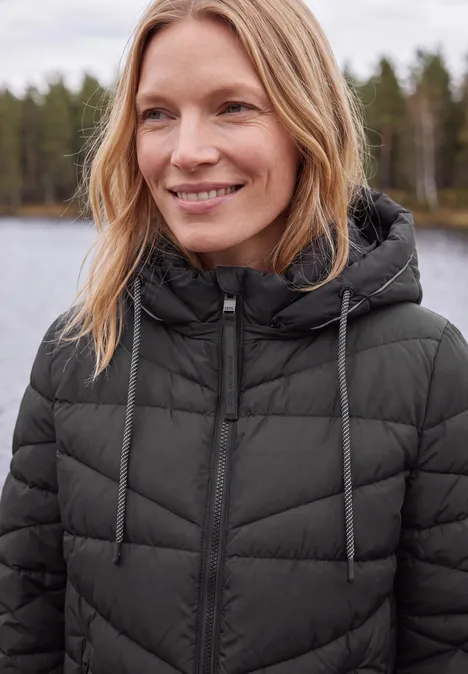Blonde Frau in schwarzer Steppjacke mit Kapuze, lächelt vor einem See und Wald im Hintergrund.