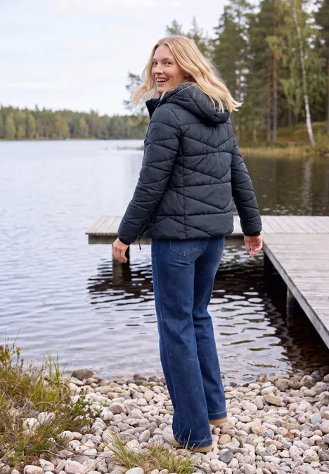 Eine Frau in schwarzer Steppjacke und Jeans lächelt am ruhigen Seeufer neben einem Holzsteg im Wald.