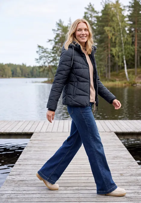 Eine Frau spaziert lächelnd auf einem Steg am See, trägt schwarze Jacke und Jeans.