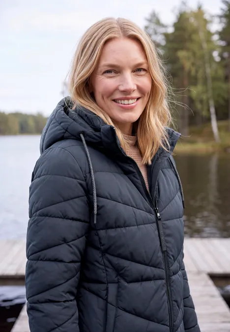 Eine lächelnde Frau in schwarzer Steppjacke steht auf einem Steg an einem ruhigen Waldsee.