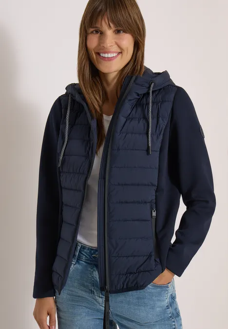 Eine lächelnde Frau in dunkelblauer, gesteppter Kapuzenjacke, weißem Shirt und Jeans.