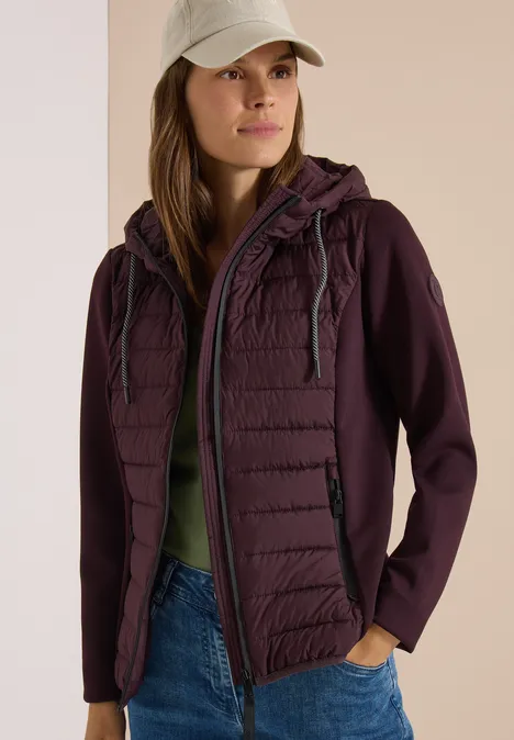 Femme portant une veste matelassée violette à capuche, jeans et casquette devant un fond clair.