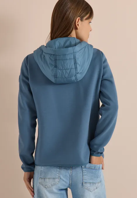 Frau in blauer Kapuzenjacke mit gesteppter Kapuze und Jeans, Rückansicht vor neutralem Hintergrund.