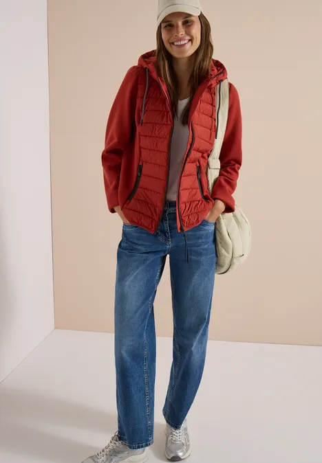 Frau mit roter Kapuzenjacke, roter Steppweste, blauen Jeans, weißen Sneakers und Cap vor beigem Hintergrund.