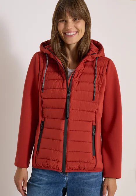 Eine Frau in roter gesteppter Kapuzenjacke und Jeans lächelt vor hellem Hintergrund.