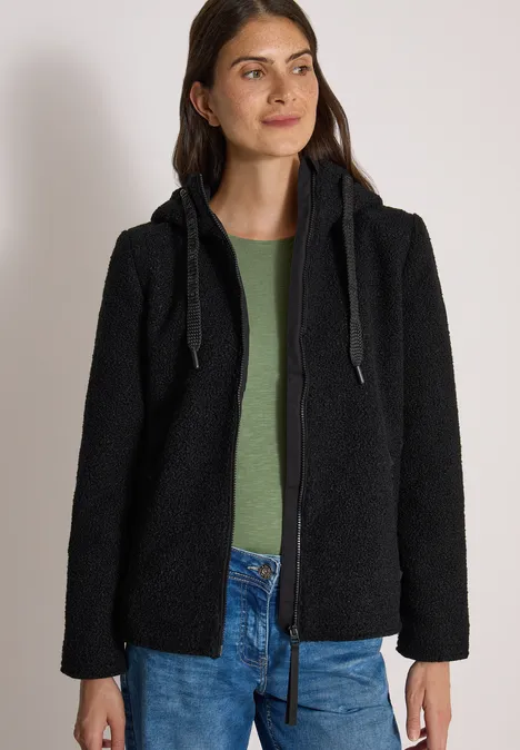 Frau trägt schwarze Teddy-Fleecejacke mit Kapuze, grünes Shirt und blaue Jeans.