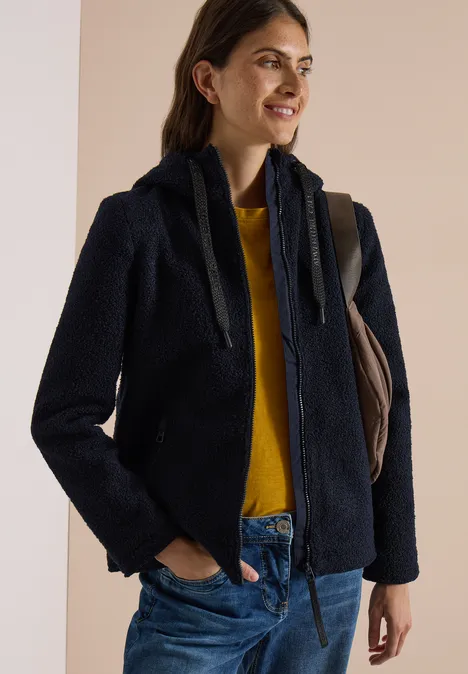 Eine lächelnde Frau trägt eine marineblaue Teddyjacke, gelben Pullover und Jeans, mit Schultertasche.