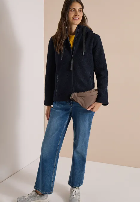 Frau in dunklem Wollmantel, Jeans und gelbem Pullover, hält eine braune Clutch vor hellem Hintergrund.