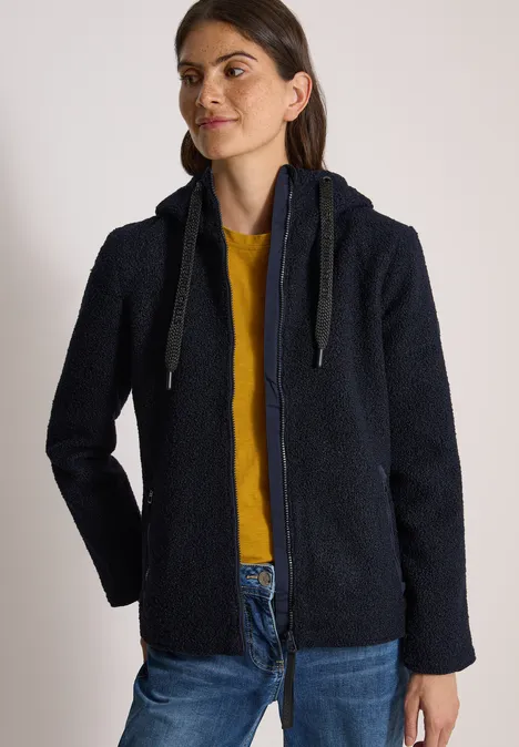 Frau im dunkelblauen Teddy-Fleece mit Kapuze, gelbem Shirt und Jeans.