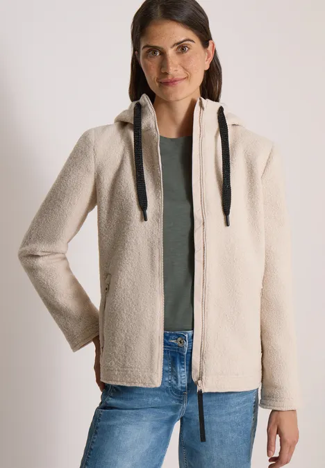 Frau trägt cremefarbene Teddy-Fleecejacke mit Kapuze, grünes Shirt und Jeans.