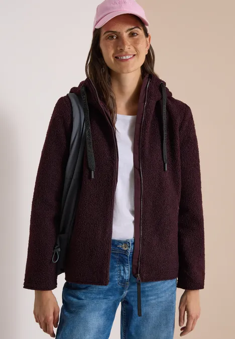Frau in dunkelroter Teddy-Fleecejacke, weißem T-Shirt, Jeans und rosa Cap, mit Rucksack und Lächeln.