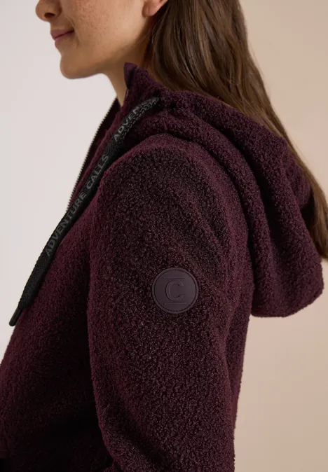 Frau trägt eine lila Teddy-Fleecejacke mit Kapuze, Reißverschluss und Markenlogo am Ärmel.