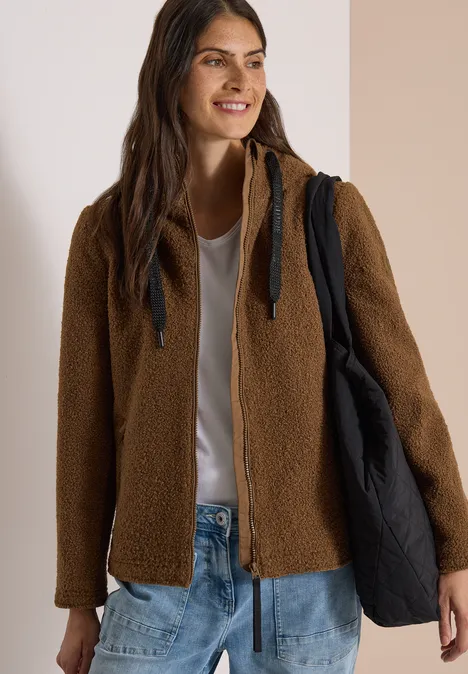 Eine Frau trägt eine braune Teddyjacke, weißen Shirt, Jeans und schwarzen Shopper, sie lächelt.
